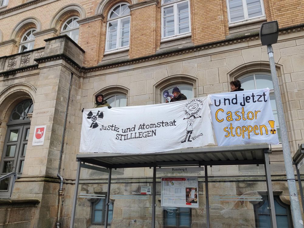 Menschen stehen vor dem Landgericht auf dem Dach einer Bushaltestelle mit Banner: Justtiz- und Atomstaat stilllegen und jetzt Castor stoppen! mit Atom-totenkopfzeichen