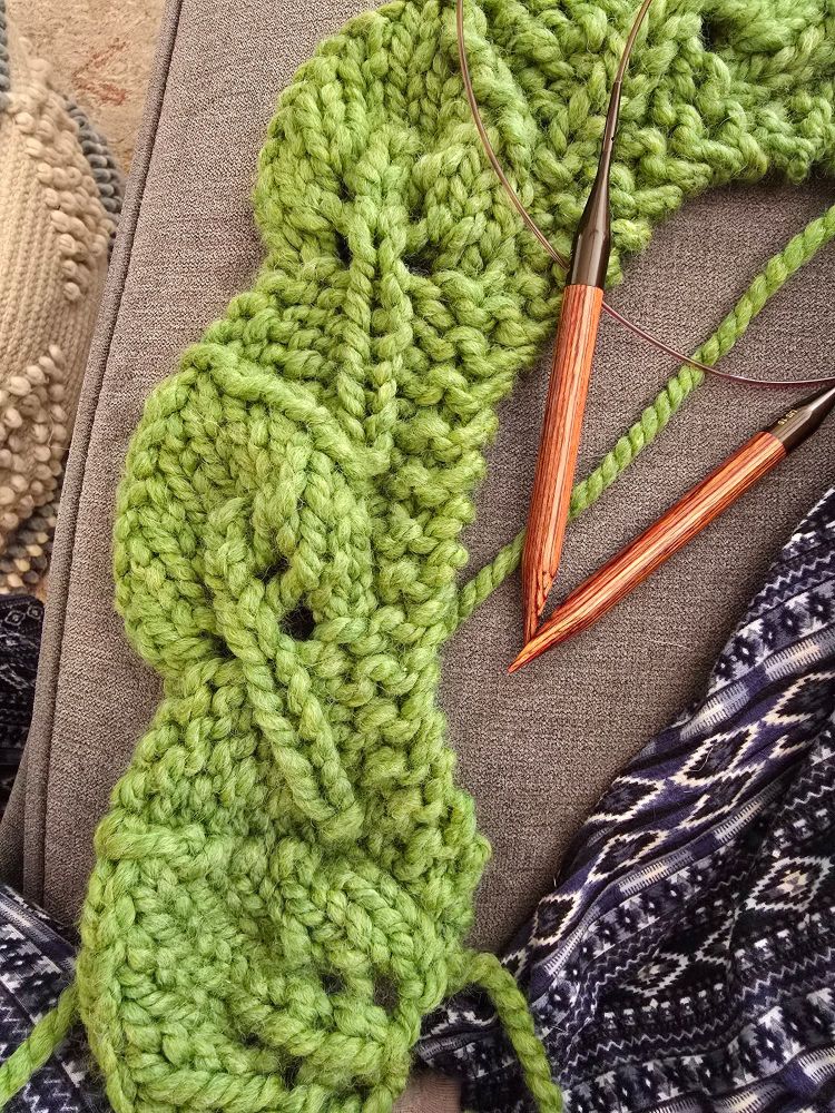 A green scarf in the process of being knitted is made of a repeating leaf design.