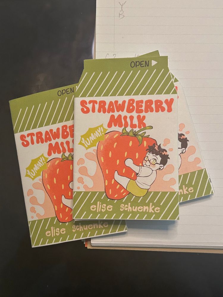 Three mini zines in the shape of milk cartons lay on paper and a screen. the cover has a green and red color palette with the title: STRAWBERRY MILK with a label of YUMMY

a dark haired person wearing a white shirt and light green shorts smiles while holding onto a giant strawberry that splashes up pink milk