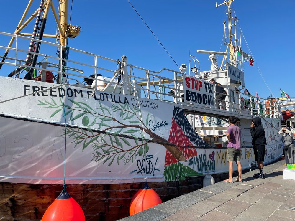 This is a photo of the Freedom Flotilla boat. It is covered in different illustrations and messages and has a sign that says stop the genocide. 