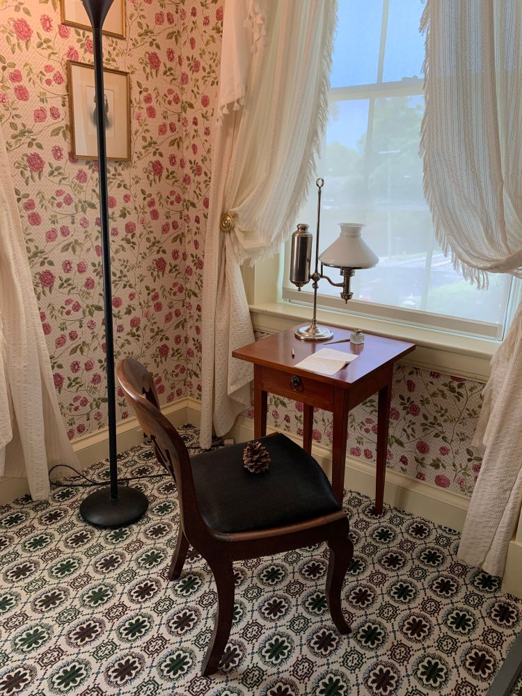 In a Victorian bedroom, a very small table the size of a side table and a regular size chair are near the window. The table is set with a small lamp, a sheet of paper, an inkwell, and a pen.