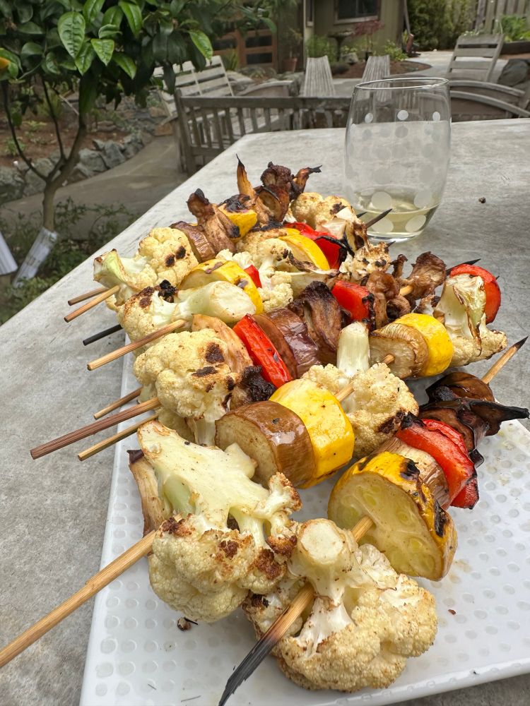 Several skewers laying diagonally on a white plate, with a glass of white wine in the background, on a counter outdoors 