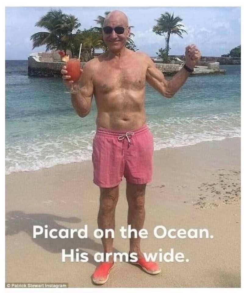 Sir Patrick Stewart on a tropical beach in pink swim trunks holding a tasty looking frozen beverage aloft