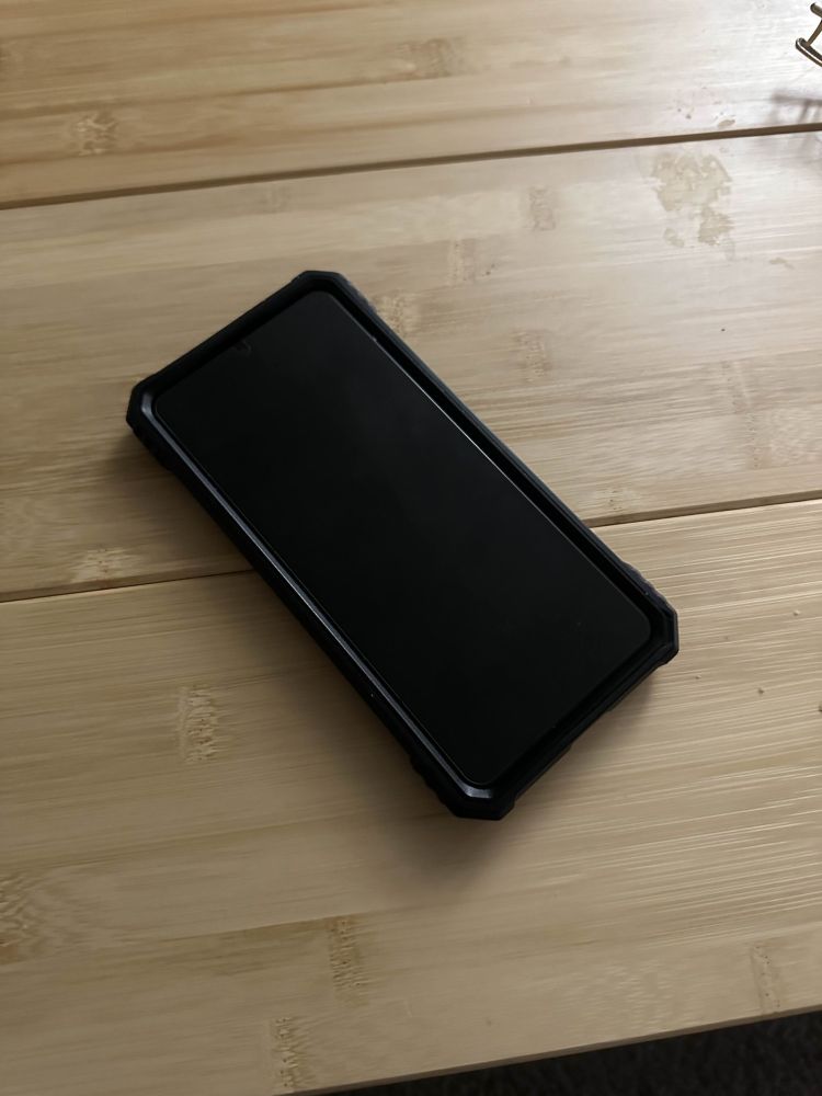 A Samsung Galaxy S21 Plus placed on a wooden table. The phone is turned on and functioning despite the screen being completely blank with no backlight.