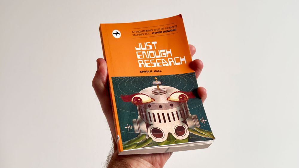 A hand holding a book titled Just Enough Research by Erika Hall, featuring a retro sci-fi cover design