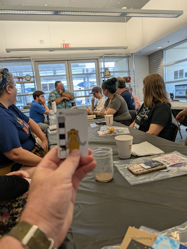 Hand holds a scale bar with a small mastodon on it, in front of a long table of paleontologists