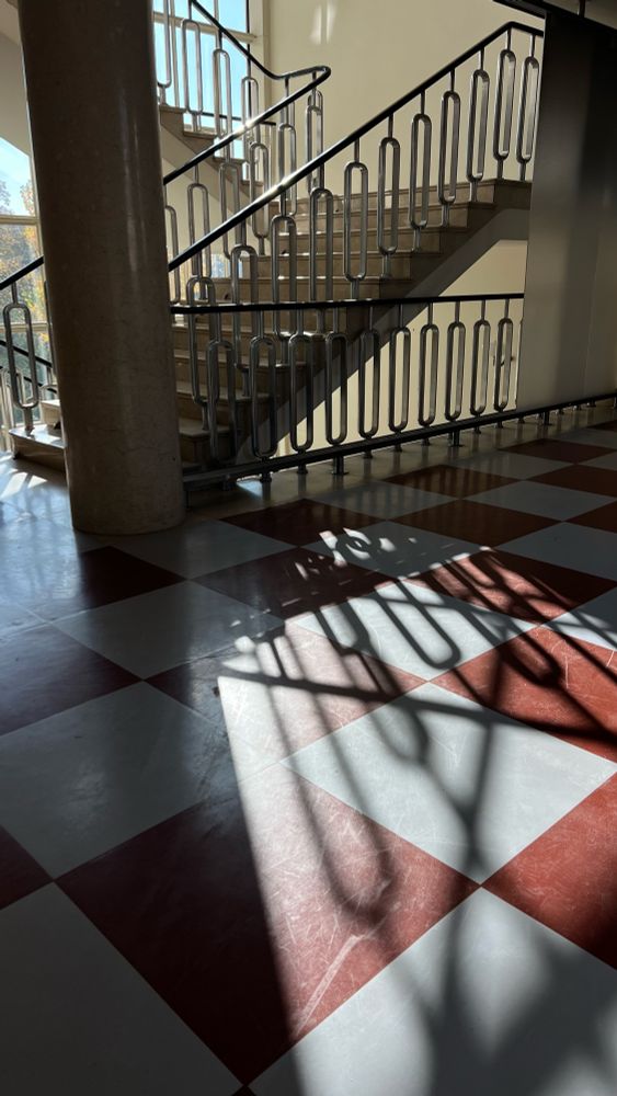 Ein Treppenhaus mit modernistischer Architektur, geprägt von großen Glasfenstern, die viel Tageslicht hereinlassen. Die Treppe hat ein elegantes Geländer mit geometrischen Formen und führt an einer glänzenden schwarzen Säule vorbei. Der Boden ist aus beigem Stein und erzeugt zusammen mit den klaren Linien der Treppe eine stilvolle, lichtdurchflutete Atmosphäre.