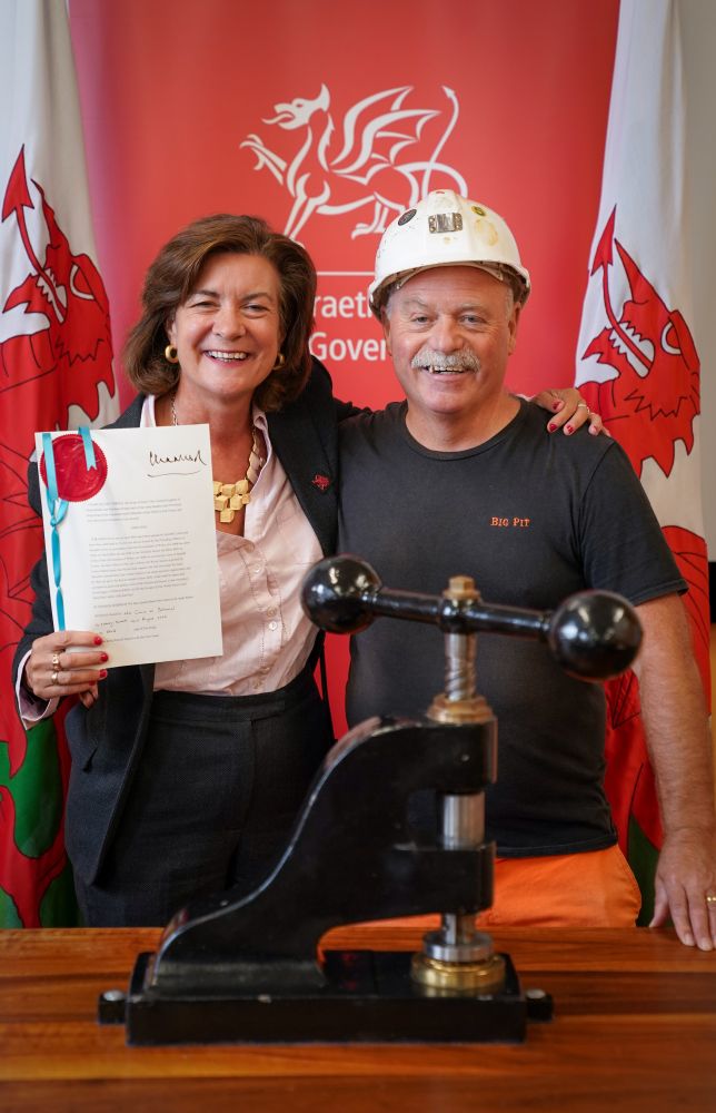 The First Minister of Wales smiling and standing next to a man wearing a white hard hat and a "Big Pit" T-shirt during a bill sealing ceremony at Big Pit. The First Minister is holding up a signed bill with a red wax seal and blue ribbon. Behind them are two Welsh flags and a red backdrop featuring the Welsh Government logo.