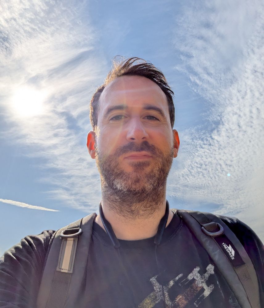 Me looking down on the camera with the sun shining in the backdrop. I'm wearing a rainbow Star Wars tee and a rucksack. There are some clouds in the sky but it is a glorious day. 