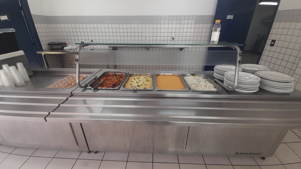 Balcão de almoço com pratos brancos, arroz, polenta, macarrão ao molho branco, frango ensopado e doce de leite de sobremesa.