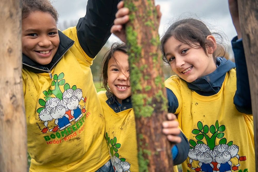 Close-up beeld van kinderen die een boom in de grond zetten. Ze kijken lachend in de camera, met gele Boomfeestdag-shirts aan.