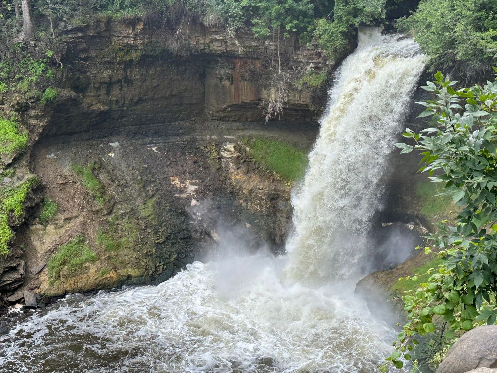 A waterfall with heavy, fast flow, kicking up plumes of water when it lands in the small pool eroded out of layers of sandstone 