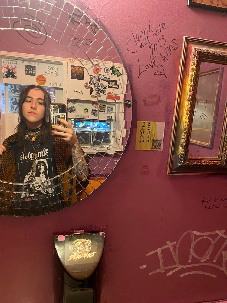 A mirror selfie of myself in the venue bathroom. I am wearing an amygdala band shirt, an assortment of necklaces, and a brown keffiyeh.
