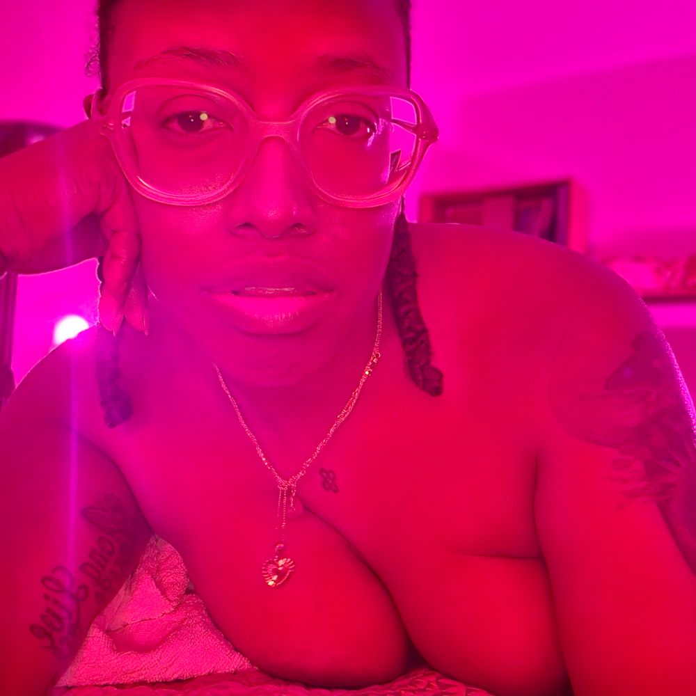 Black femme with glasses and 2 braids looks into camera. They're lying on their stomach, top of titties and some tattoos showing. Pink light bathes everything in the picture pink.