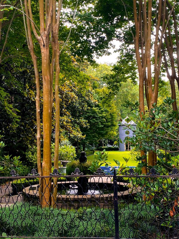A beautifully landscaped yard of a home in Charleston, South Carolina has a circle shaped water fountain with a statue in the middle tucked between tall trees and shrubs. A well maintained yard with trimmed hedges and some sort of tall structure are in the distance. A detailed cast iron fence divides the property from the public street. 