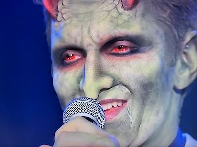 The green skinned and tiny horned demon Lorne (Andy Hallet) holding a microphone to his face. He has a blue shirt and some color jacket on. From Angel Season 2, Episode 1.