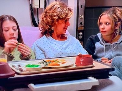 Pictured from left to right Dawn Summers (Michelle Trachtenberg), Joyce Summers (Kristine Sutherland), and Buffy Summers (Sarah Michelle Gellar). Joyce is in a hospital gown with dots. Dawn is staring at some gelatin she is picking at while wearing a green sweater with a zipper. Buffy is wearing earrings and a navy blue and grey hoodie and grey sweatpants. Dawn has brown hair while Joyce and Buffy are blonde. There is a tray of food in the foreground. Dawn and Joyce are in the latter's hospital bed. Buffy is huddled in a chair. There is medical equipment and electrical sockets in the background. From Season 5, Episode 9 of Buffy The Vampire Slayer.