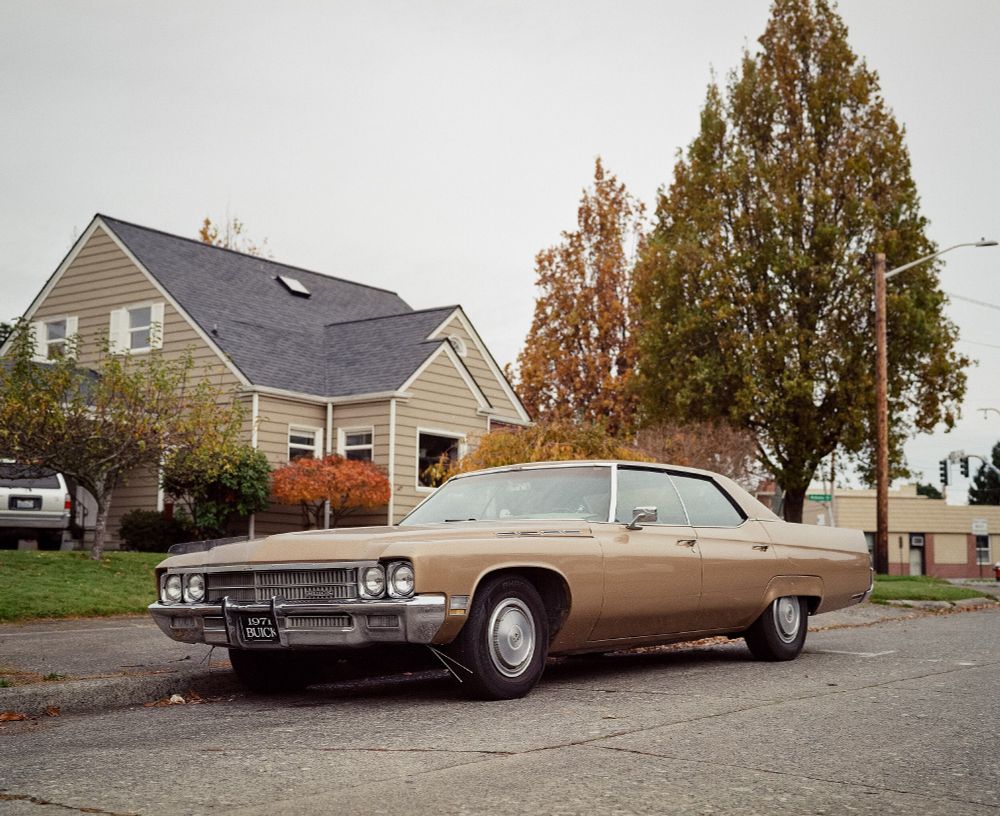 A stock 1971 Cornet Gold Buick sedan is parked on the street in a neighborhood with the chrome curb feelers gently touching the curb. Photo taken from a low three-quarter view on an overcast day. The front license plate proudly says "1971 Buick," a clear bug guard is mounted to the hood, and all of the chrome accents are shining in the subdued light.