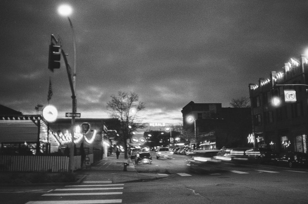 Looking down State Street at night. Heavy clouds in the sky, blurred tail lights of cars moving in the foreground, the neon sign on the Herold newspaper building is shown in the distance. Heavy grain is prominent throughout this hand-held film image. 