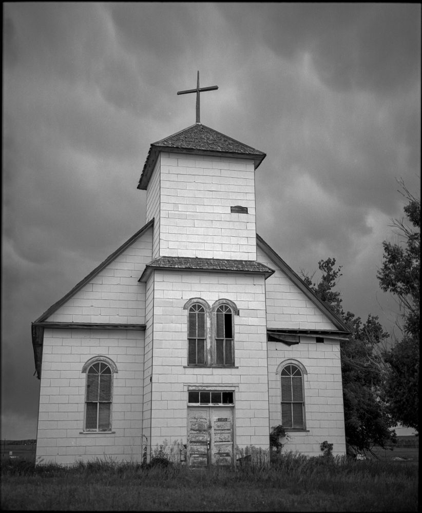 The front of St. Agnes Catholic Church in Matheson, Colorado. The dilapidated white church and steeple stand out against ominous, stormy clouds in this black and white photo. 