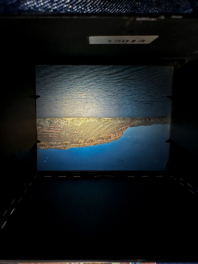 Looking through the dark cloth at the upside-down image shown on the ground glass of a Crown Graphic 4x5. The image is of Watson Lake. 