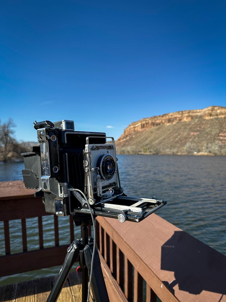 A three-quarter view of a Crown Graphic 4x5 camera on a sunny day. Watson Lake is in the background. 