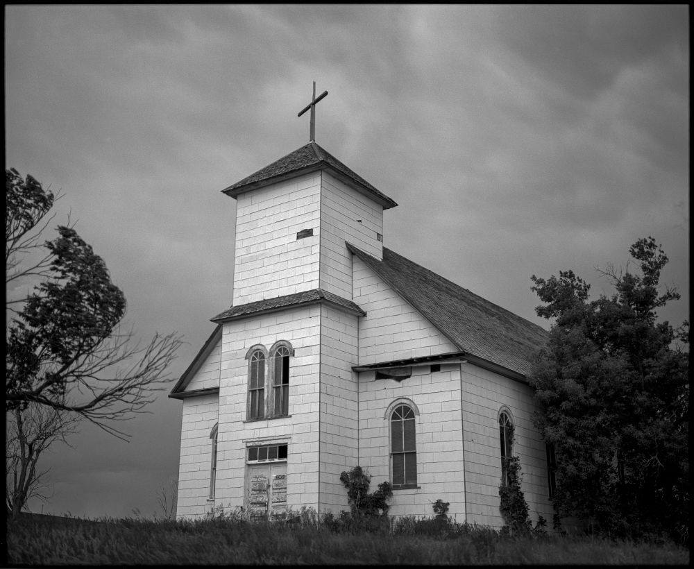 The front of St. Agnes Catholic Church in Matheson, Colorado. The dilapidated white church and steeple stand out against ominous, stormy clouds in this black and white photo. 