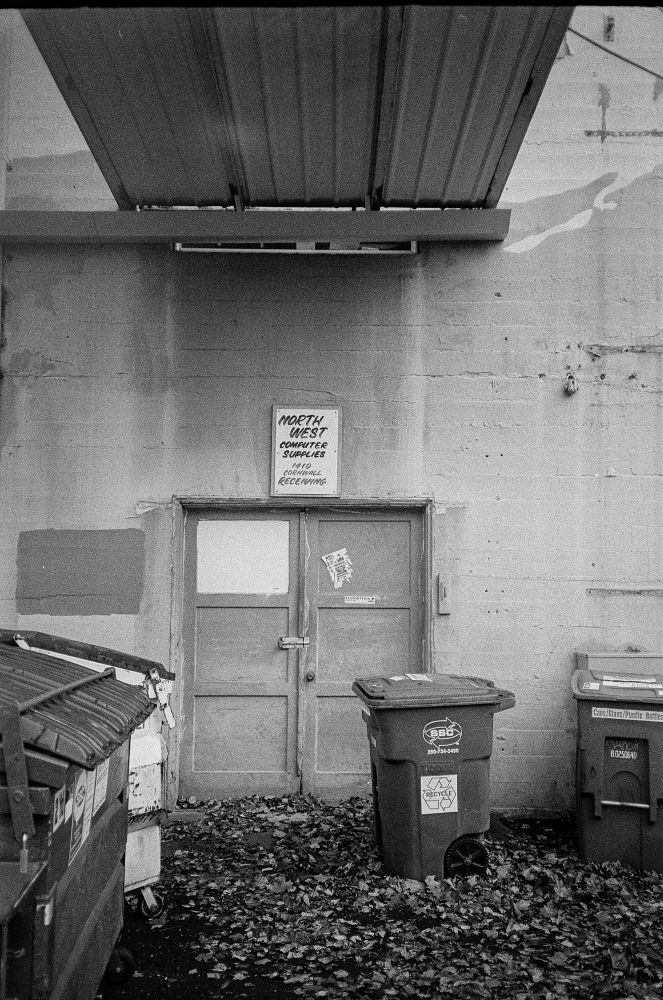 An alley, with dumpsters, garbage carts and the ground full of leaves. A hand painted sign over the doors reads “NORTH WEST COMPUTER SUPPLIES”