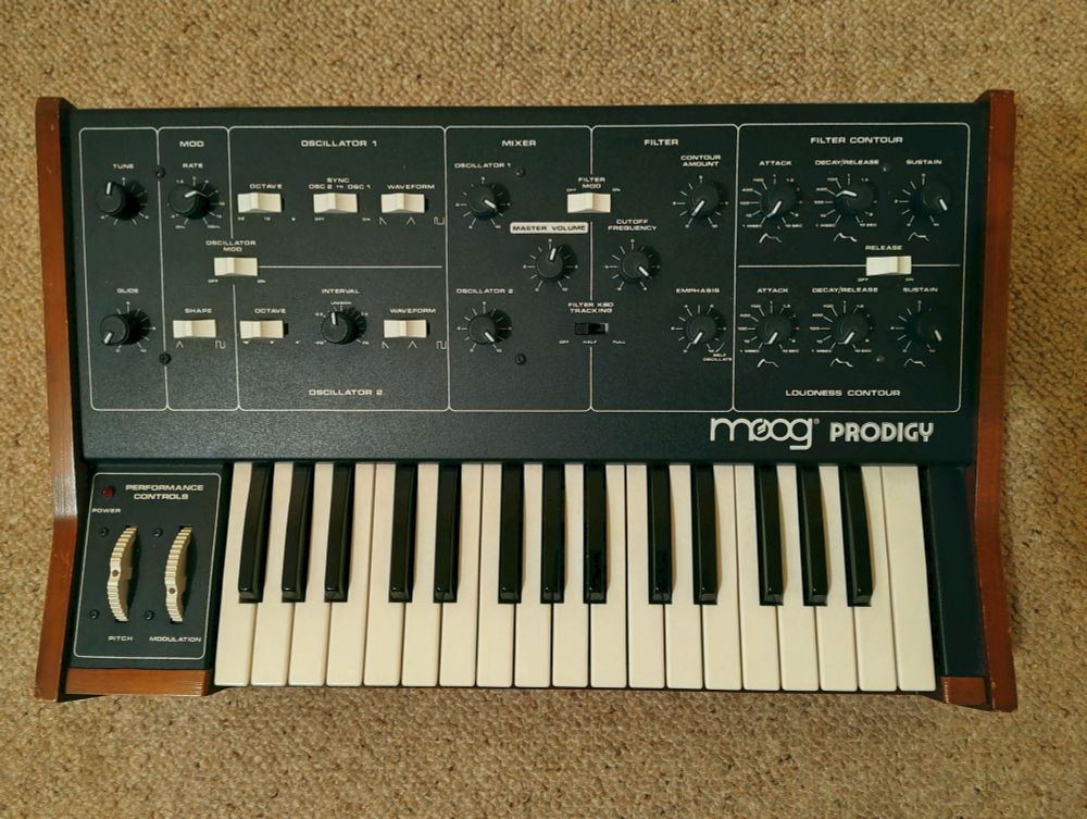 A Moog Prodigy synthesizer with a wooden casing, featuring a keyboard with white and black keys. The control panel includes various knobs and switches for sound modulation and synthesis.