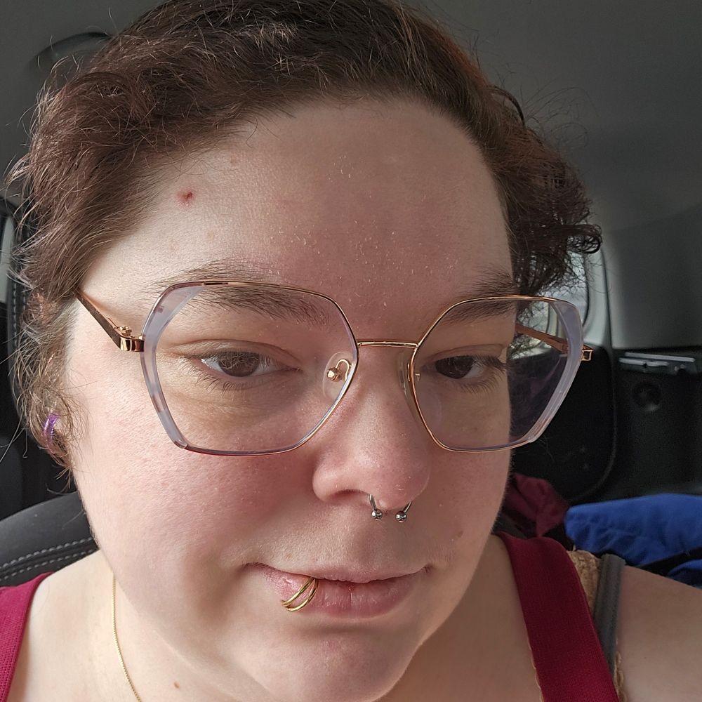 Picture of a woman with hexagon glasses, short hair and two facial piercings. Septum piercing is fresh.