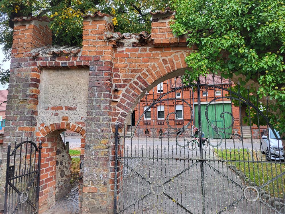 Kleine geöffnete schmiedeeiserne in einem gemauerten Tor. Daneben ein geschlossenes schmiedeeisernes Tor in einem gemauerten Rundbogen. Dahinter ein Backsteinhaus mit grünem Tor.