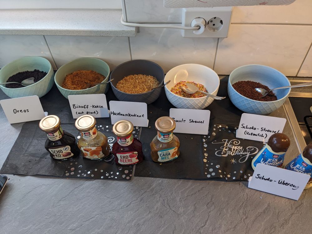 Verschiedene Schüsseln stehen auf einer Arbeitsplatte. Sie sind gefüllt mit verschiedneen Toppings und vor ihnen stehen Buffetschilder. Es sind zu sehen: Oreo-Kekskrümel, Biscoff-Kekskrümel, Haselnusskrokant, bunte Streusel und Schokostreusel. Im Vordergrund stehen noch Schokosauce, Caramelsauce, Erdbeere, Caramelsauce und Schokoüberzug.