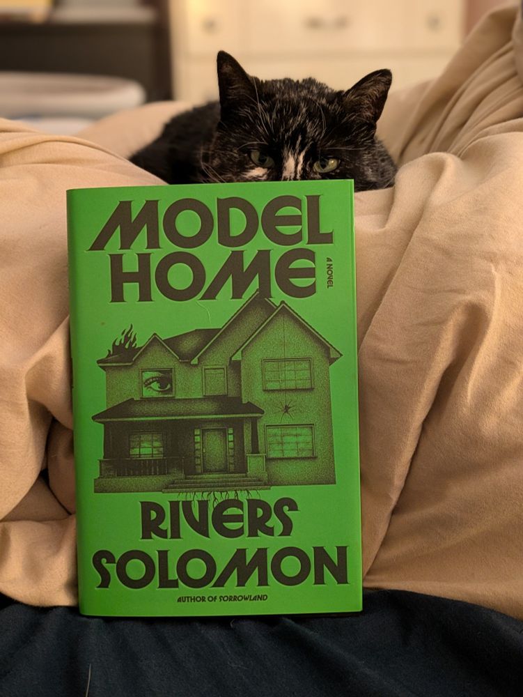 Kizzy the tuxedo tortie cat stares over the bright green book Model Home by Rivers Solomon