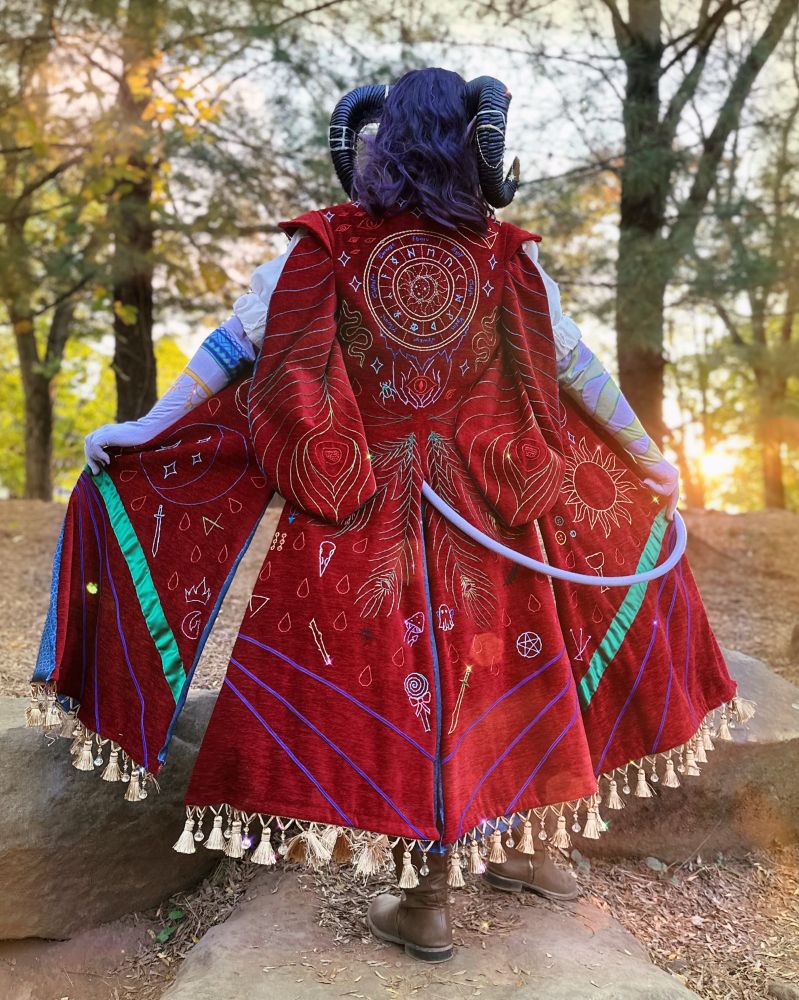 Nyx cosplaying Mollymauk, a shot from behind, showing off the embroidery in the coat. They have a dark curled purple wig, dark ram horns covered in jewelry, and purple skin with vibrant tattoos. The coat is a deep red chenille covered in hand embroidery of various symbols such as peacock feathers, celestial, blood drops, red eyes, and stars. There is a light gold tassel trim adorning the bottom of the coat.