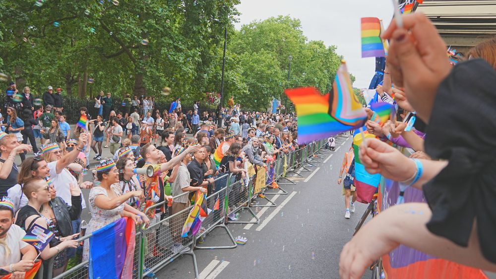 A street lined with joyful people waving colourful flags and wearing rainbow hats, cheering the parade as it goes by. 