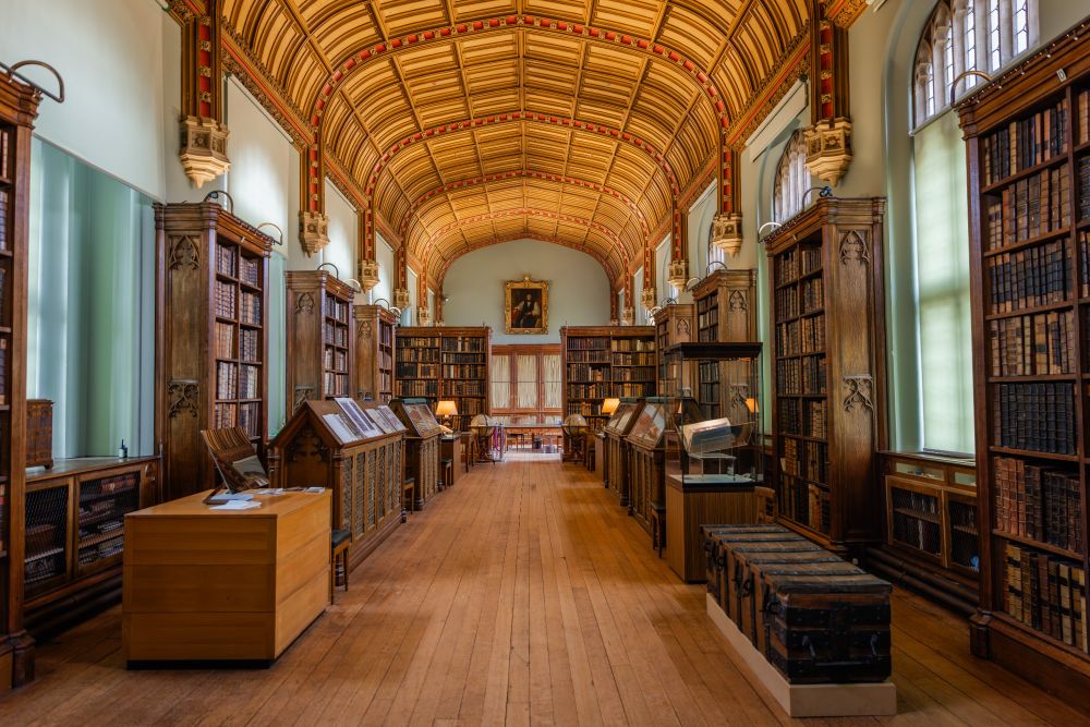 Interior of the Parker Library at Corpus Christi College in Cambridge, showcasing rows of wooden bookcases filled with books, a vaulted ceiling, and historical artifacts on display.