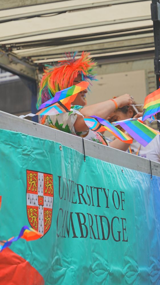 A person wearing a rainbow wig waves from a section of a London Pride parade float decorated with Progress flag bunting. The University of Cambridge logo is displayed prominently.