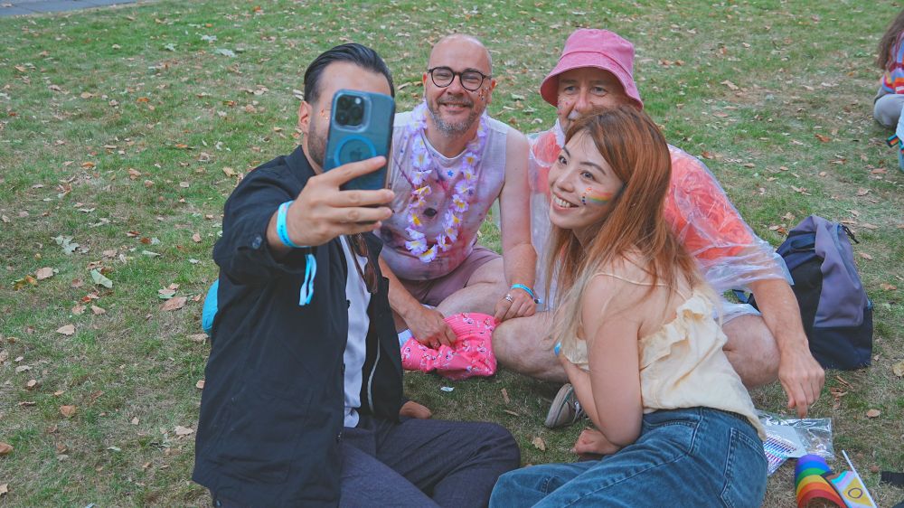 Four people sitting on a grass patch taking a group selfie. They're dressed colourfully and are wearing glittery face paint.