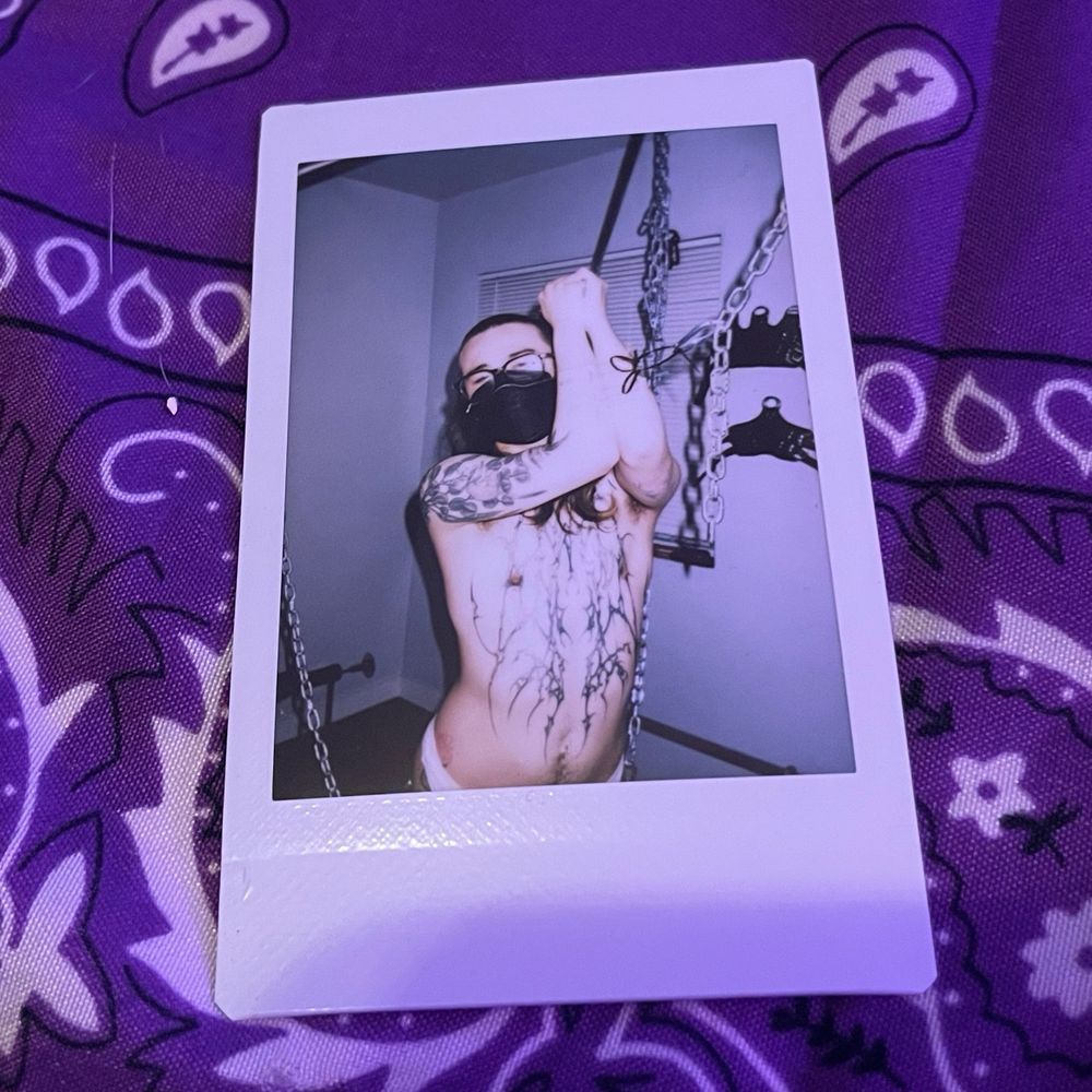 polaroid of boy mischief from the waist up. it is wearing a black mask, its needle bound arms held above its face, suspended with zipties through the needles, linked to chains 