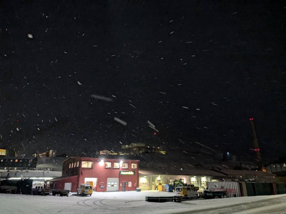An almost pitch black snowy scene, with artificial lighting from a small town catching on falling snow