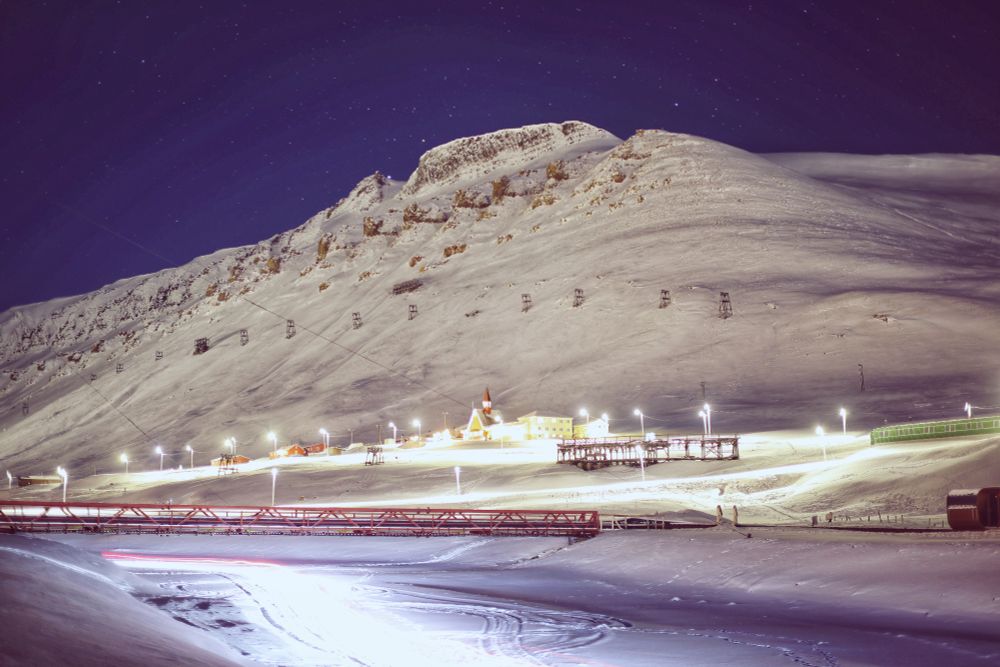 A snow covered mountain with a church, lit by streetlights, under a purplish sky. In the foreground, traffic trails from a snowmobile pass under a low bridge
