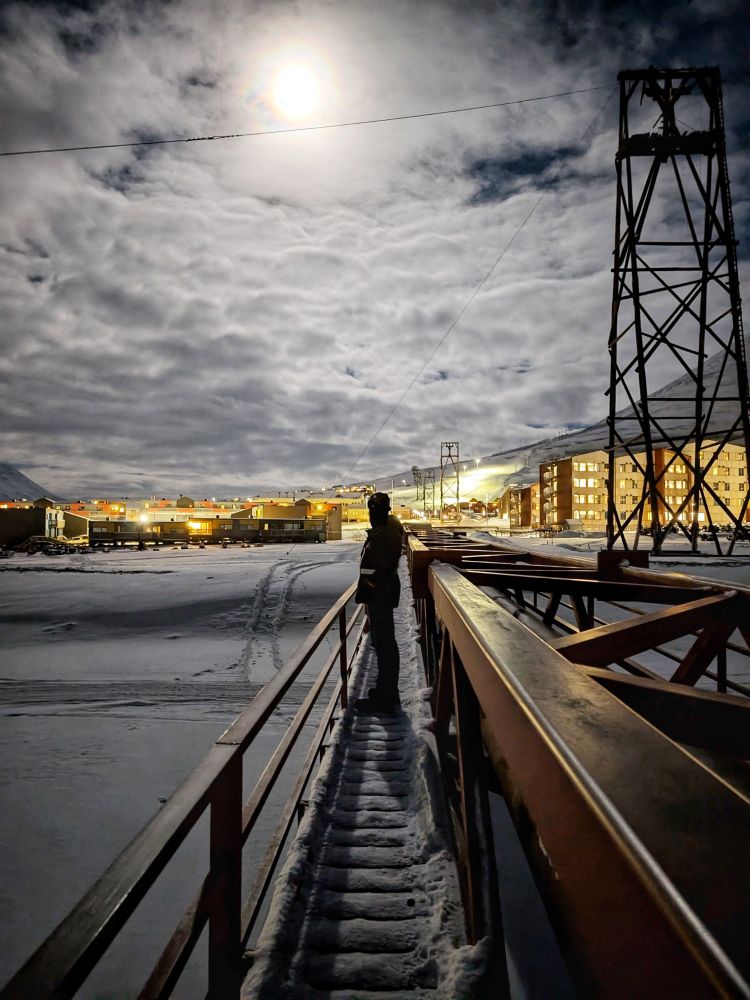 A figure stands on a snow covered bridge. It is the middle of the night but the full moon is so bright that the figure and the bridge are both casting a clear shadow
