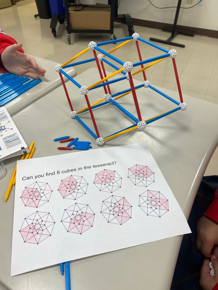 Zometool construction of a hypercube sitting on a student desk behind a sheet of paper containing 8 Schlegel diagrams of a hypercube.  The student has shaded the location of 8 different cubes, one for each diagram.  