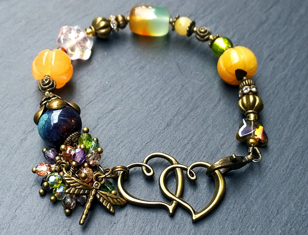 My new handmade Enchanted Garden bracelet. It has antiqued brass double hearts and dragonfly charm, jade pumpkins, cluster of fire polished glass dangles, agate and ceramic beads. It has orange tones with blues and greens. 