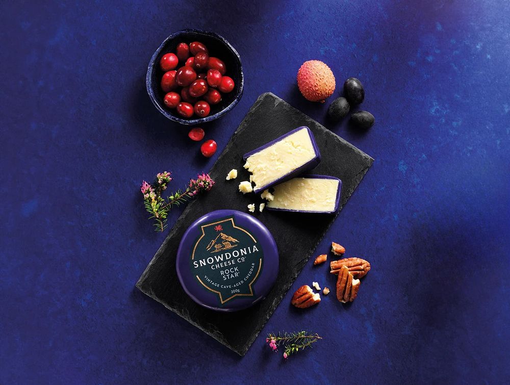 A cutting board against a deep blue background filled with tasty cheddar cheese, nuts, berries and other snacks.