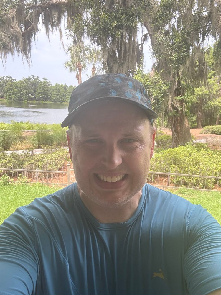 Selfie with sloth running hat, blue shirt, and pond in background. 