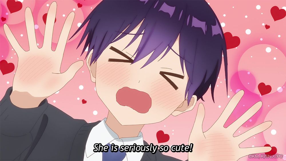 A close-up of Izumi from Shikimori's Not Just a Cutie with his hands raised, blushing, and surrounded by hearts with text underneath that reads, 