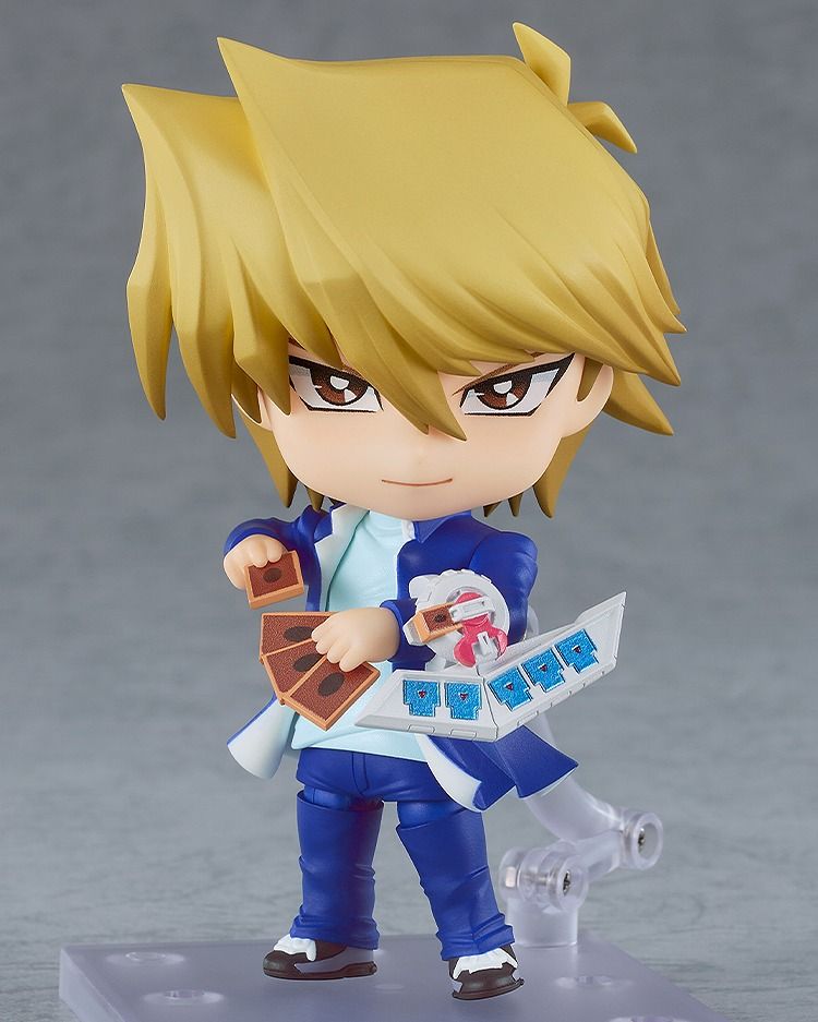 A picutre of Yu-Gi-Oh! - Joey Wheeler Nendoroid with his Smiling face. On one arm is his  Duel Disk, and on his other hand he is holding cards.
