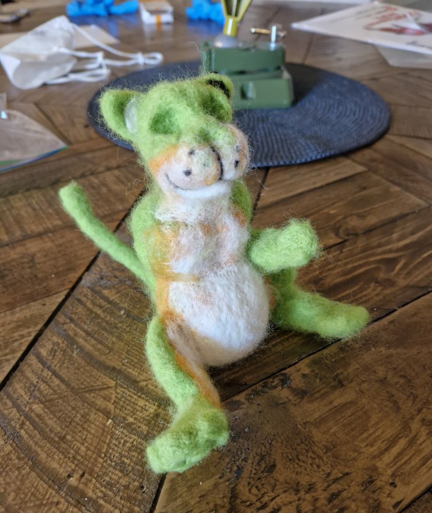 an unfinished needle felted figure of a green cat with a white and orange belly. he  has two legs, a tail, one arm and no nose or eyes 