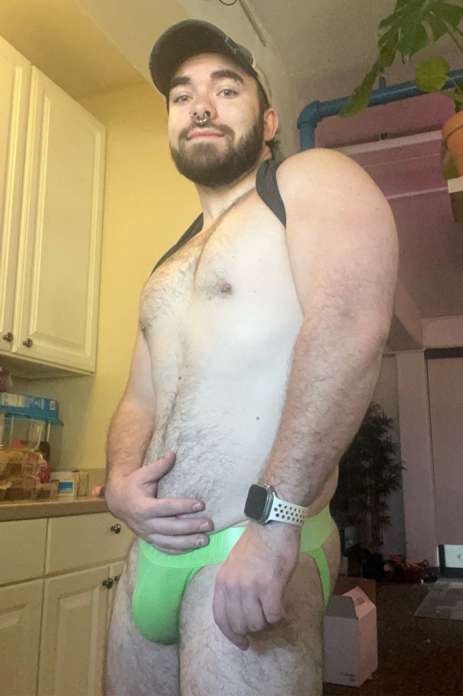 Mostly nude photograph with person posed with their hand placed on their stomach in a green jockstrap.